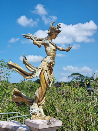 Golden statue of a dancing woman with flowing robes, set on a stone railing. Lush green foliage and a blue sky form the background.
