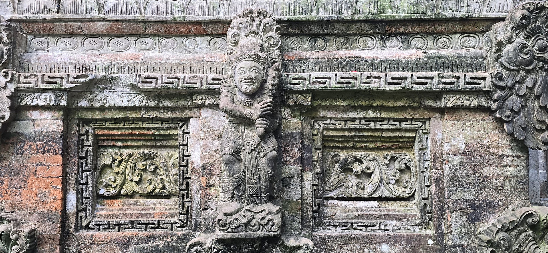 Ornate weathered stone temple wall with carved guardian figure and floral panels in gray and mossy green tones.