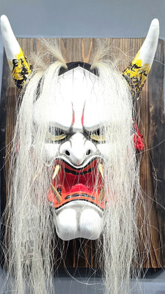 A traditional white mask with fierce expression, horns painted yellow, and long hair on a wooden background, conveying an intense mood.