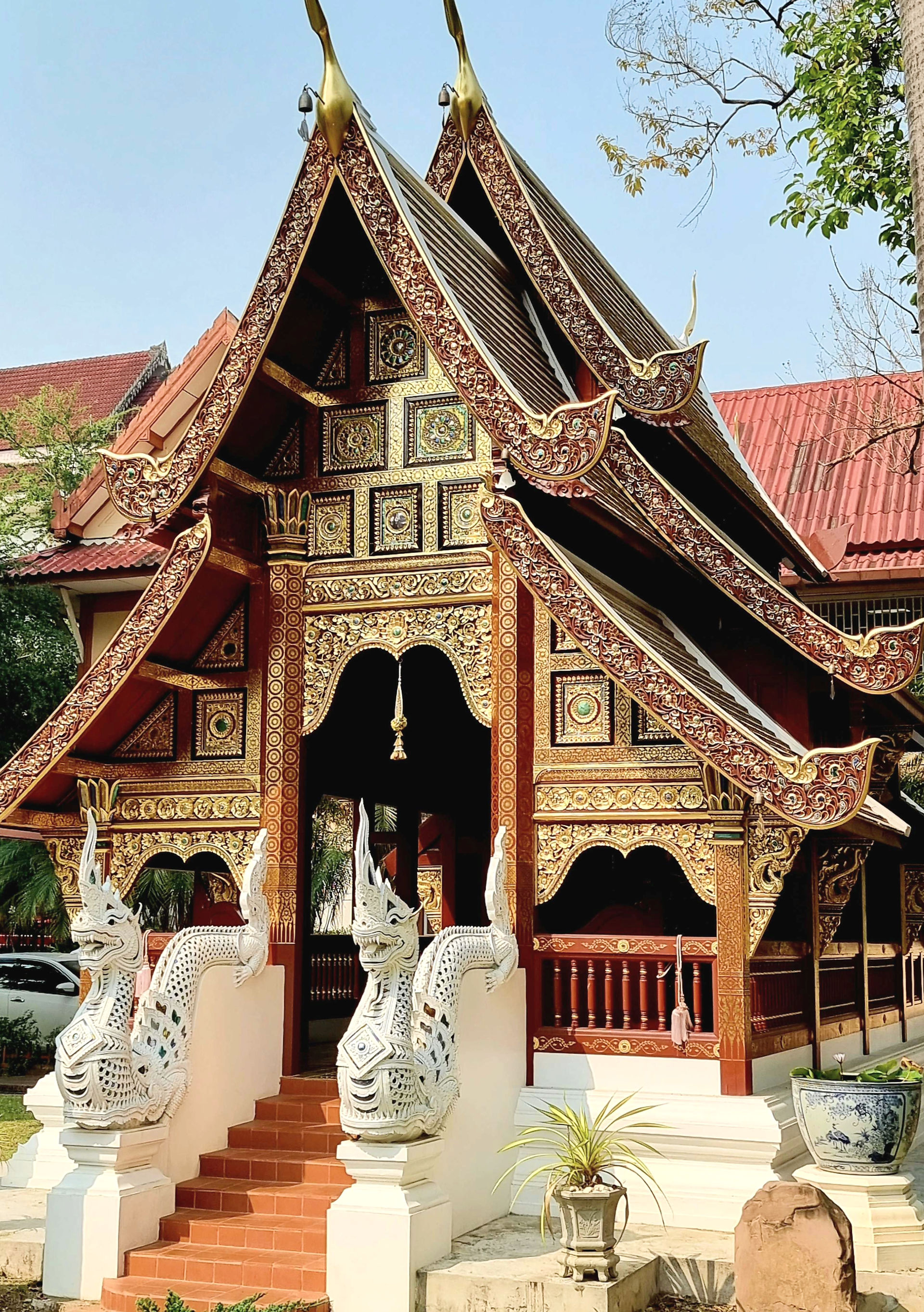 ornate timber virharn covered in gold engravings with 2 pointed roofs, 2 white naga guards along a small, red brick staircase