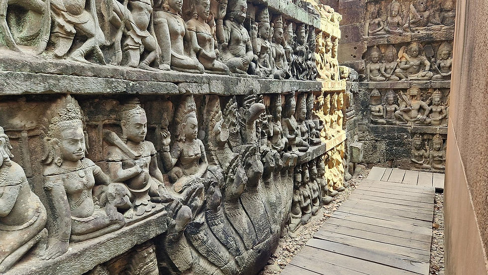 Stone carvings of seated figures on temple walls, featuring intricate details, line a narrow wooden walkway. The scene is peaceful and ancient.