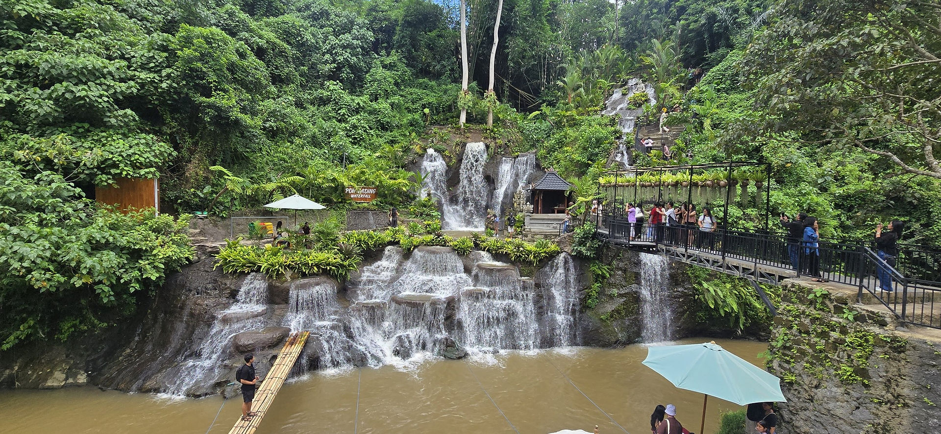 People enjoy a lush green waterfall setting, with a bamboo bridge and viewing platform.