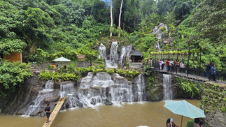People enjoy a lush green waterfall setting, with a bamboo bridge and viewing platform.