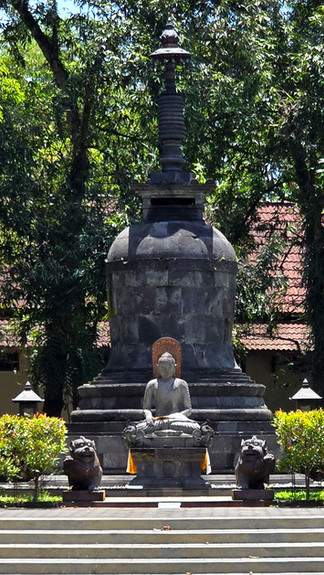 Stone Buddha statue in a garden, seated in meditation before a stupa. Green trees in the background, flanked by guardian figures.