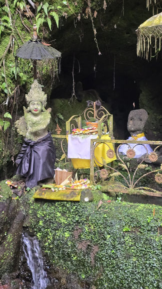 Mossy cave shrine with Balinese-style statues, yellow and black drapes, flower offerings, and a small waterfall in lush greenery