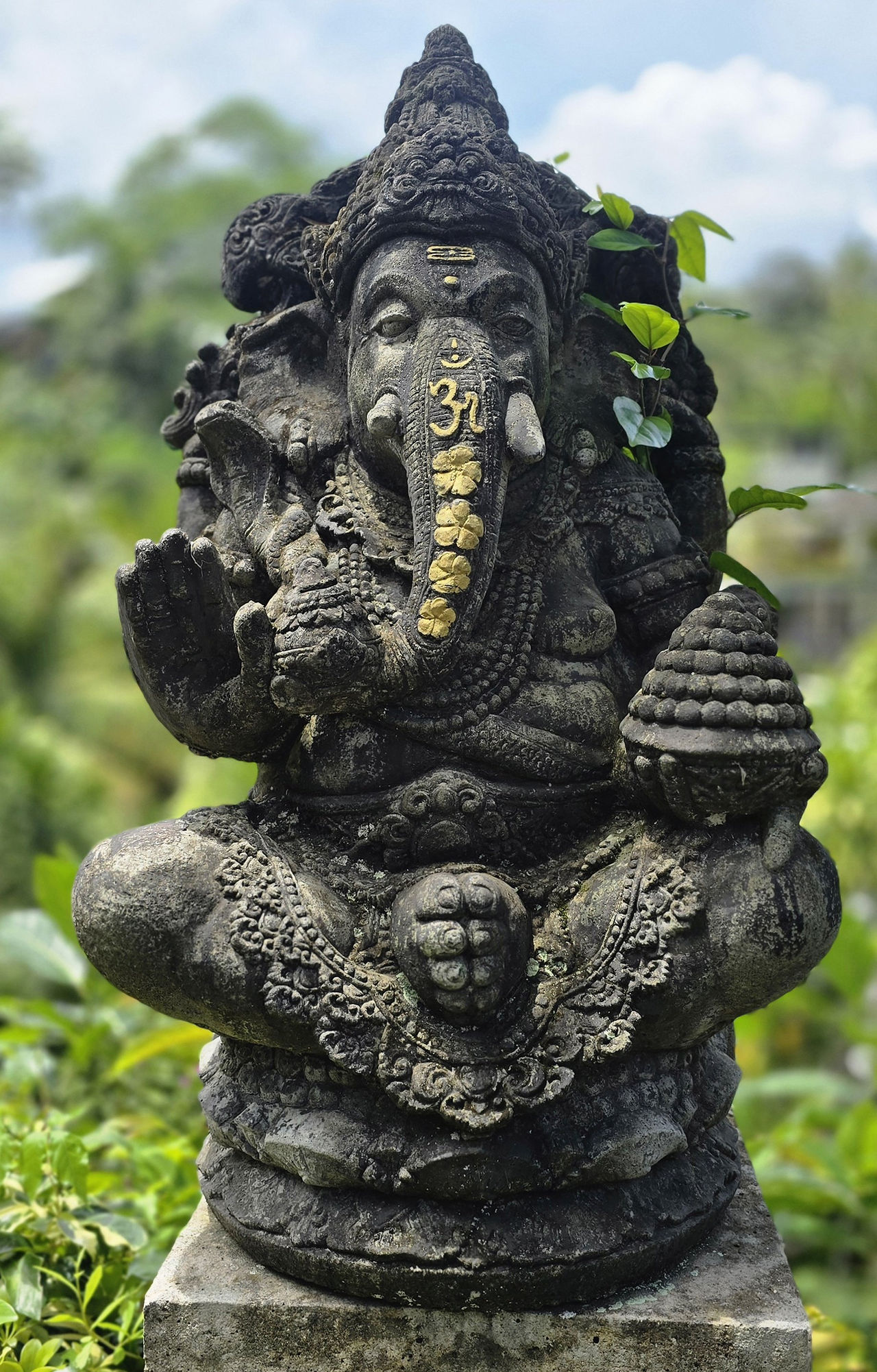 Black stone ganesha statue with gold symbols on its trunk, leafy green jungle in the background 