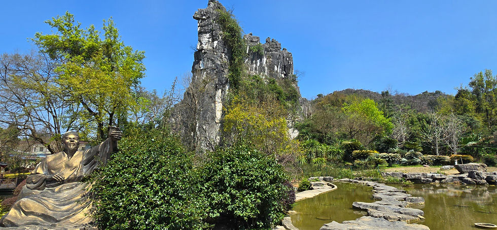 Golden statue of a seated figure holding a cup, surrounded by lush greenery and trees, with a rocky hill in the background under a clear blue sky.