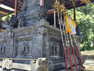Ornate stone temple with red and white poles leaned against it, intricate carvings, and gold accents. Lush greenery in the background.