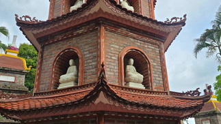 Pagoda with intricate roof tiles, featuring seated Buddha statues in niches. Surrounded by lush trees, under a cloudy sky.