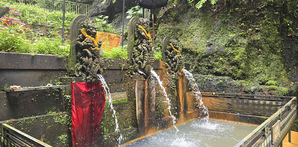 Stone statues with ornate carvings spout water into a pool, surrounded by lush greenery. Red fabric and orange garlands accent the scene.