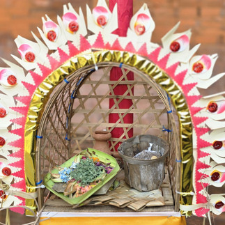 Decorative woven altar with pink-white flower border, tray of offerings and metal bucket, against a brick wall.