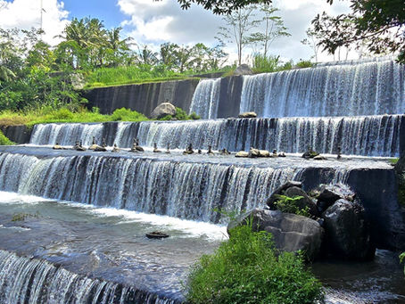 Tiered waterfall with clear water flowing over stone ledges, surrounded by lush greenery and a bright blue sky, creating a serene scene.