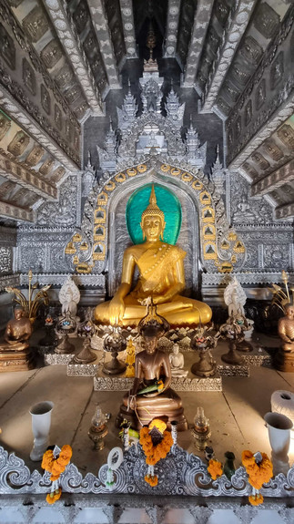 Golden Buddha statue in ornate temple, surrounded by smaller statues and vibrant marigold garlands. Intricate silver details adorn the space.