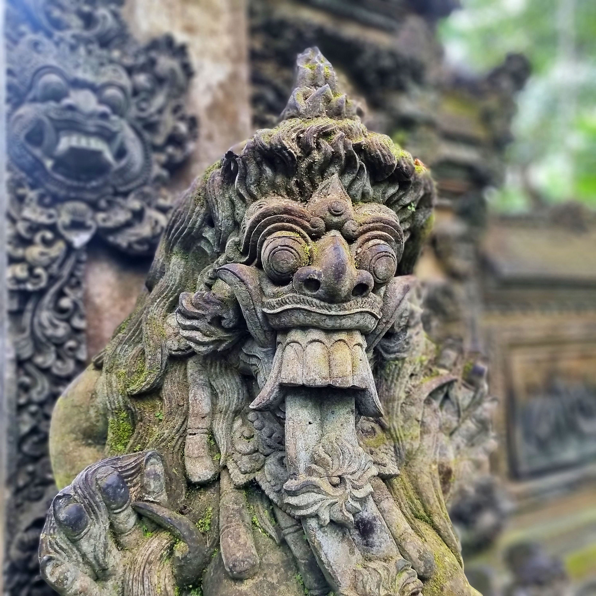 Stone statue of a fierce mythological creature with intricate details and mossy textures, set against an ornate, blurred background.