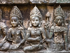 Stone carvings of three seated deities with ornate headdresses and serene expressions, set against an ancient, weathered stone wall.