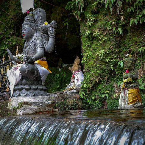 Stone Hindu statues in a mossy cave shrine beside a flowing waterfall, draped in white and yellow cloth with a serene, sacred mood