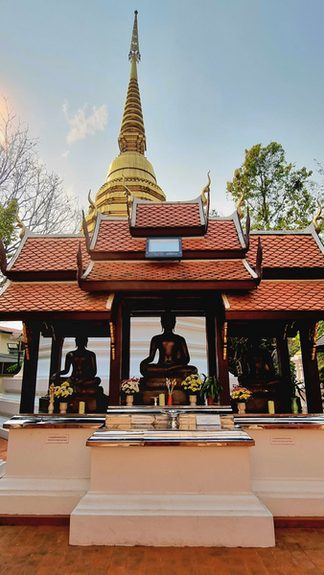 Buddha statues under a red-roofed pavilion, with a golden stupa in the background. Sunny sky, trees, and offerings visible. Peaceful ambiance.