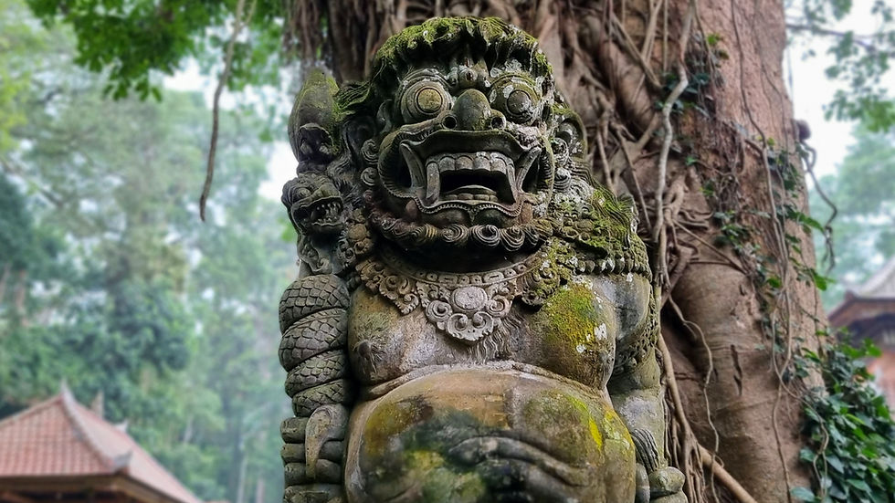 Stone statue of a fierce figure with large eyes and mouth, covered in moss, stands against a tree with vines in a lush, temple setting.