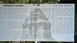 Signboard of Selogriyo Temple with English and Javanese text, detailing temple history and structure. It stands outdoors among trees.