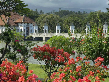 Tranquil garden scene with colourful flowers, a white bridge, and a traditional house by a pond, surrounded by lush greenery and trees.