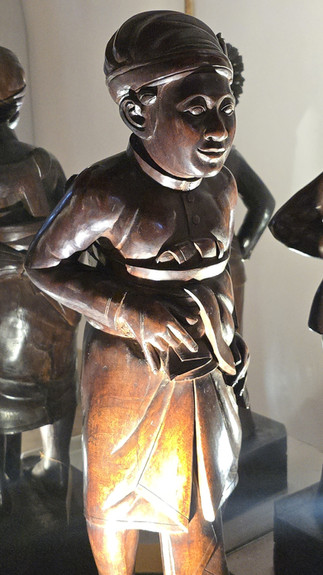 Wooden sculpture of a person with detailed features and wrapped garment, illuminated, set against a plain background. Other statues partially visible.