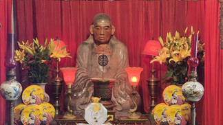 Buddhist altar with a statue surrounded by red curtains, lilies, red lamps, and decorative plates. Calm and serene atmosphere.