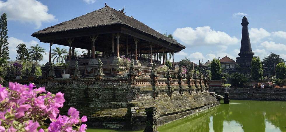 Traditional Balinese pavilion with thatched roof surrounded by stone statues, lush greenery and pink flowers, set against a clear sky.