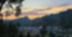 Sunset over a town with colorful skies and silhouetted mountains in the background. Trees frame the scene, creating a serene mood.