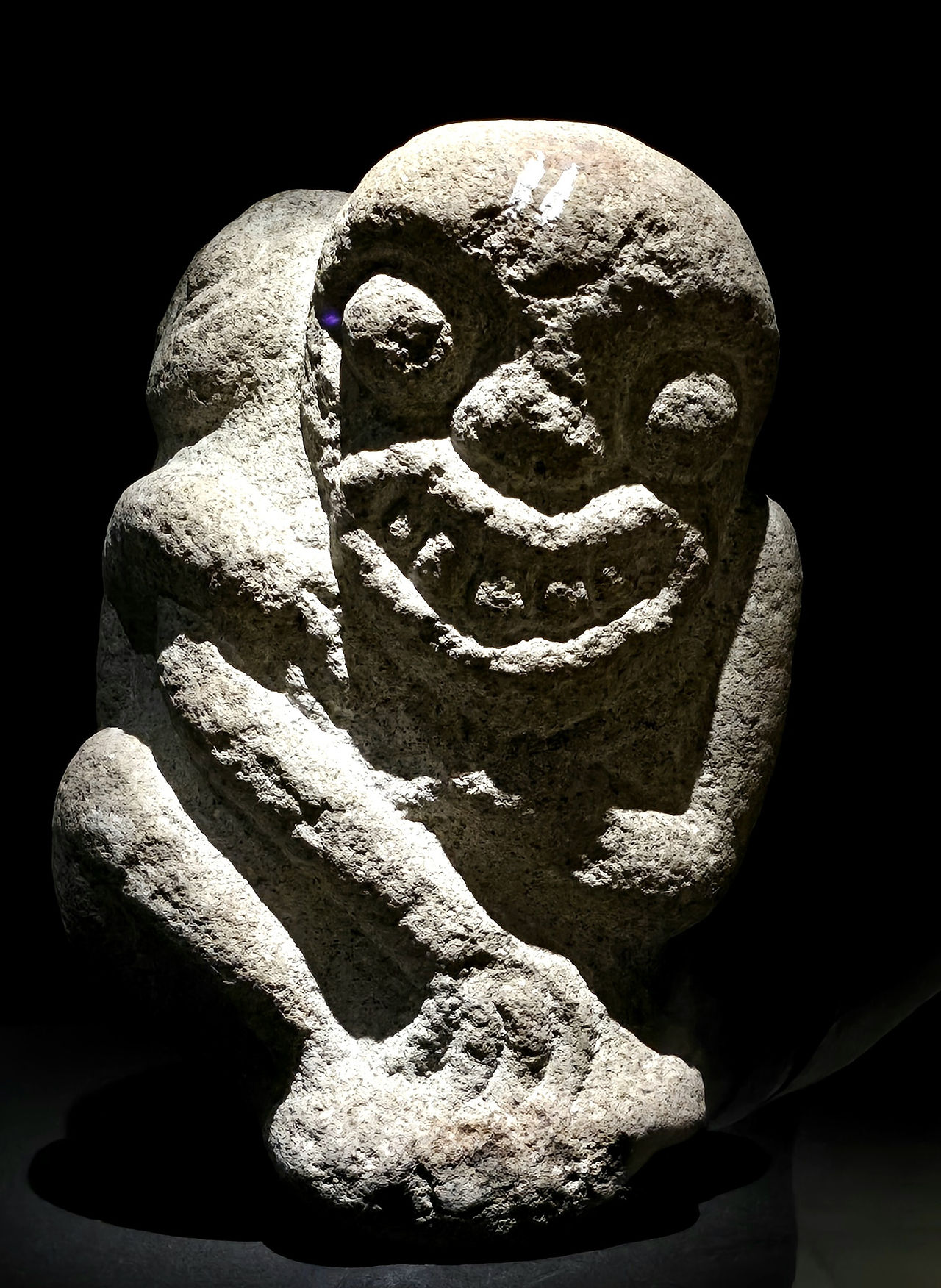 Stone statue of a grinning figure crouched, illuminated against a dark background. Rough texture and prominent facial features.