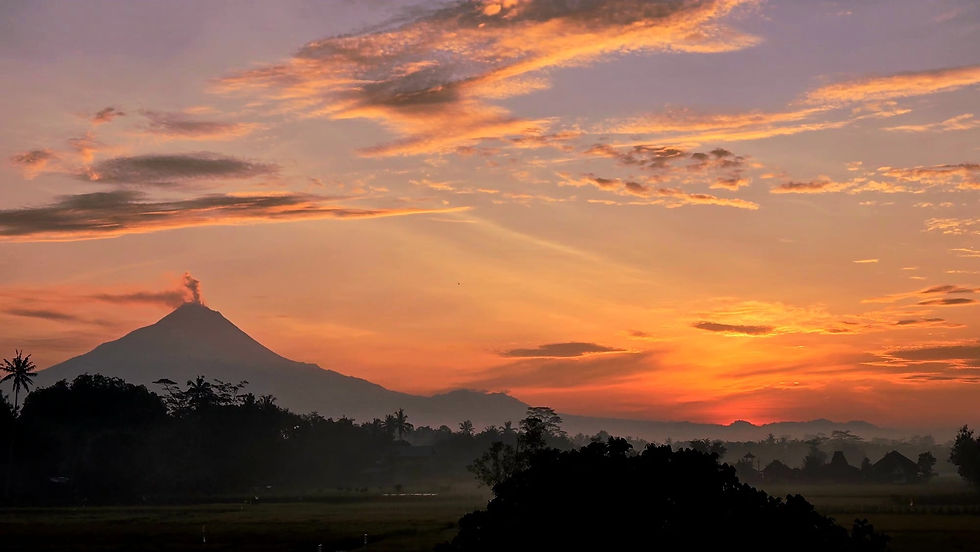 Mount Merapi v
olcano emits smoke at sunrise with a vibrant orange sky and scattered clouds. Silhouetted trees and buildings in the foreground.
