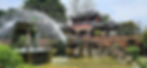 Ancient Chinese architecture with red lanterns, large bronze vessel fountain, and greenery. Water sprays gracefully in a serene setting.
