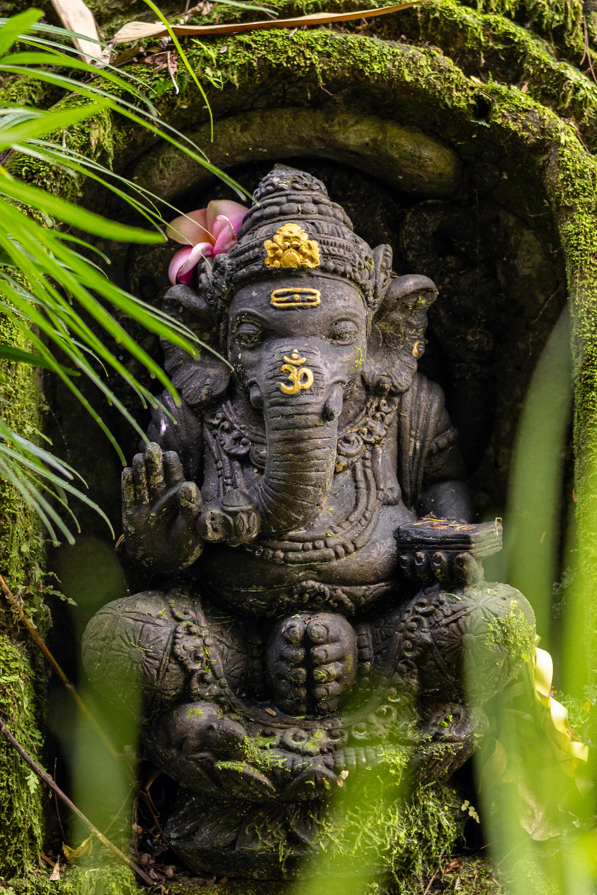 Ganesha statue in a mossy niche, adorned with pink flowers and gold details. Lush greenery surrounds, creating a serene atmosphere.