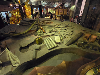 Museum exhibit with historical artifacts, including artillery shells and nets, in a dimly lit setting. Visitors in the background.