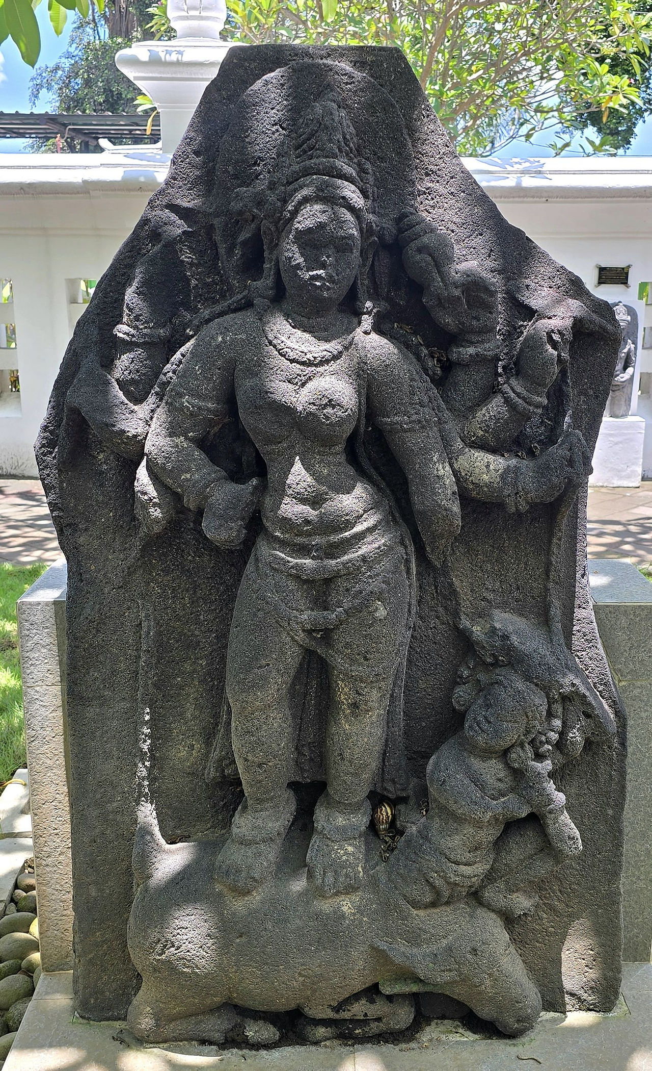 Stone sculpture depicting a woman standing with an ornate background. Intricate details on jewelry and attire. Lush greenery in the backdrop.