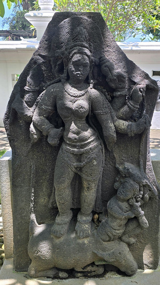 Stone sculpture depicting a woman standing with an ornate background. Intricate details on jewelry and attire. Lush greenery in the backdrop.