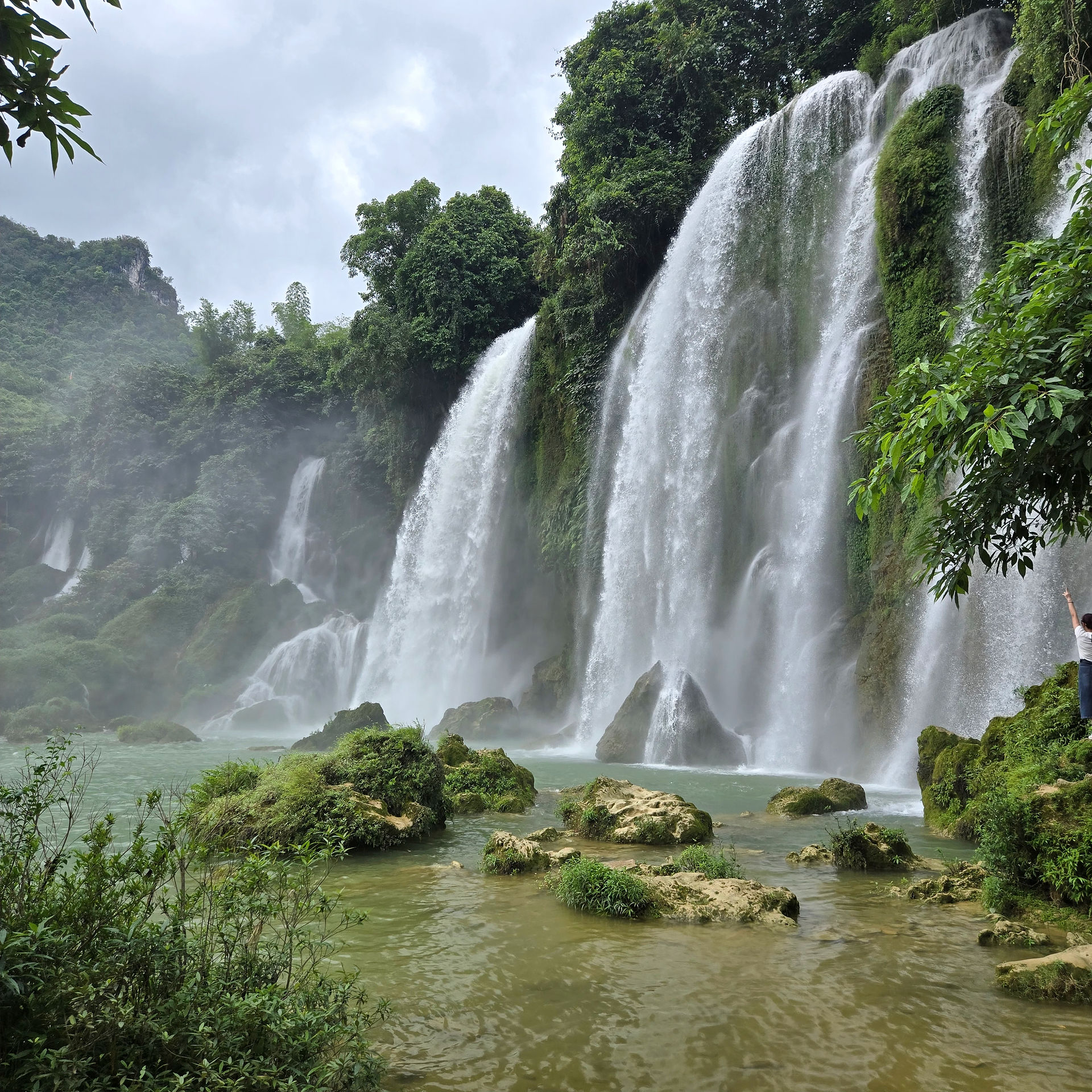 Waterfalls cascade over lush, green cliffs into a misty pool. A person on the right raises their arm. The scene is serene and vibrant.