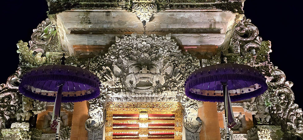 Intricate stone carvings on a Balinese temple gate illuminated at night, with two large purple umbrellas. The mood is serene and majestic.