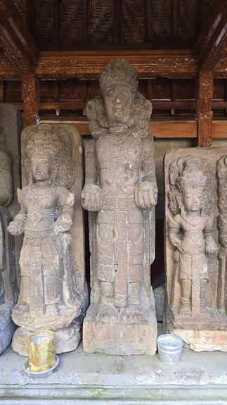 Three carved stone guardian statues stand in a wooden temple alcove, with offering cups at their feet and a solemn mood.