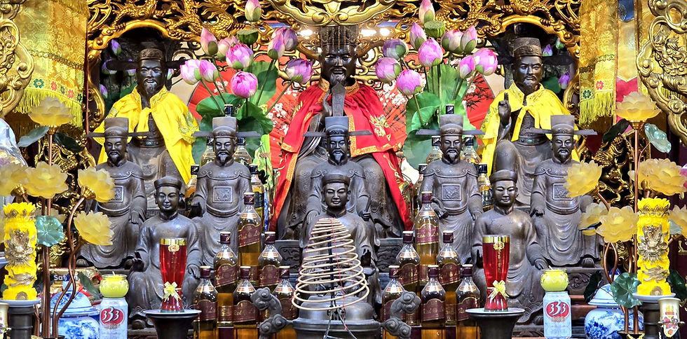 Elaborate shrine with ornate statues, red and yellow robes, vibrant pink flowers, incense coil, and Johnny Red bottles amid golden backdrop.