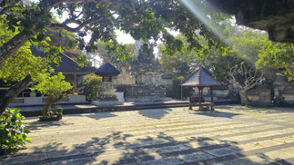 Sunlit stone courtyard with temple structures, surrounded by lush green trees and dappled shadows. Peaceful and serene atmosphere.