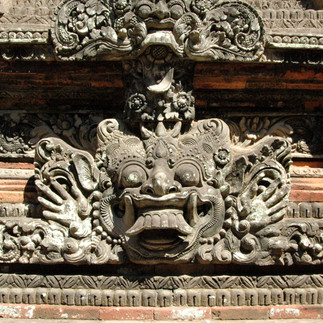 Intricate stone carving of a mythical face with wide eyes and open mouth, adorned with floral patterns, set against a brick wall background.