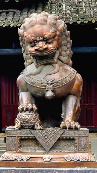 Bronze lion statue on ornate pedestal in a lush garden. Traditional Chinese building with gold accents in the background. Greenery surrounds.