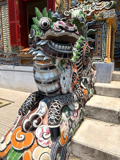 Colorful dragon sculpture with intricate patterns on steps outside a building with red shutters. Playful and vibrant atmosphere.