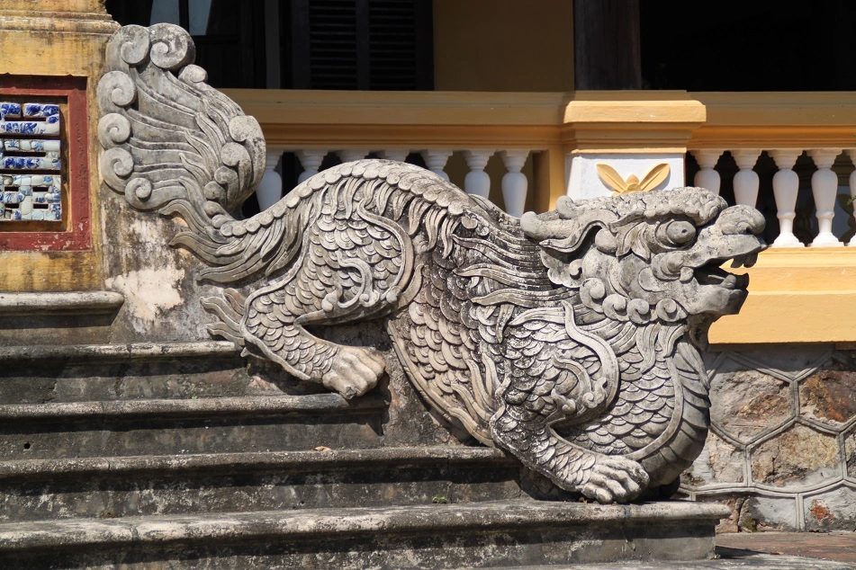 Stone dragon sculpture on staircase, adorned with intricate carvings. Yellow wall and patterned tile backdrop, evoking a historic ambiance.
