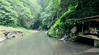 Lush jungle river with a person wading in water, a small shrine-like stall on the right, and a tiny waterfall in a calm green setting