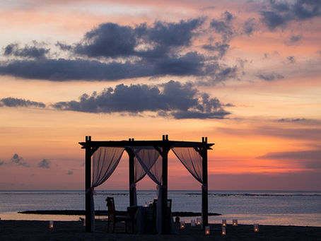 A pergola with sheer curtains on a beach at sunset, surrounded by lit candles. The sky displays pink and orange hues, creating a tranquil scene.
