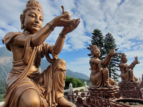 Bronze statues of figures offering objects, with trees in the background under a partly cloudy sky. Calm, serene mood.
