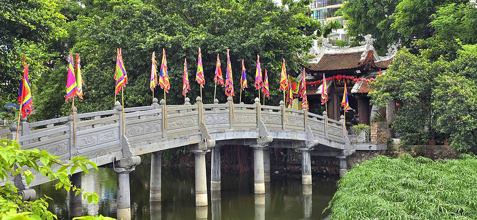 Stone bridge adorned with colourful flags crosses a pond. Lush greenery surrounds, with a traditional temple in the background under a clear sky.