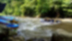 People rafting on a river in a forest, wearing yellow helmets and life jackets. Colorful rafts are stacked nearby. Lush greenery surrounds.