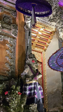 Stone statues with floral garlands stand at a temple entrance, adorned with a purple umbrella and intricate carvings, creating a serene atmosphere.